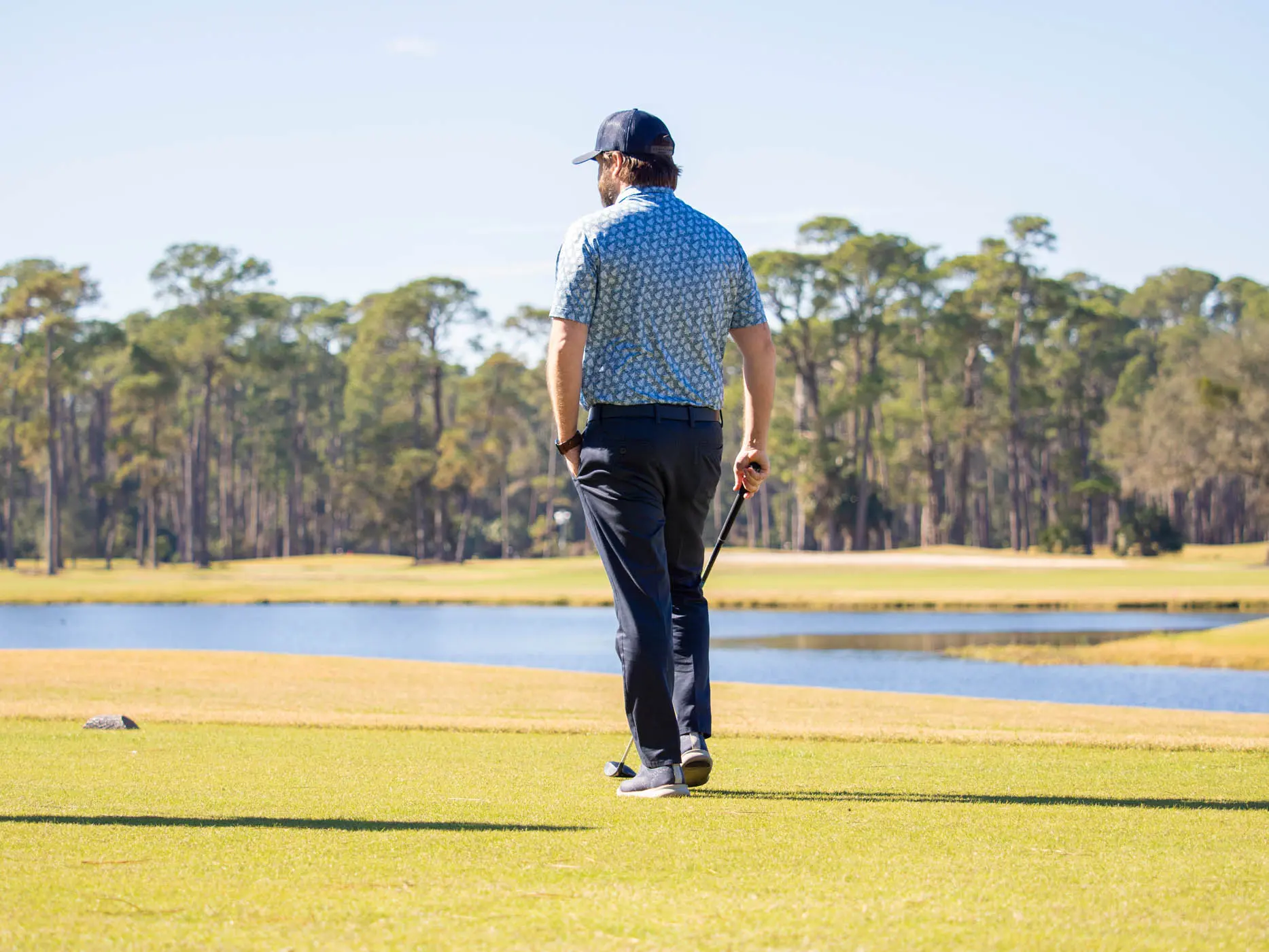 A man with a golf club in one hand and his other hand in his pocket walking on the golf green