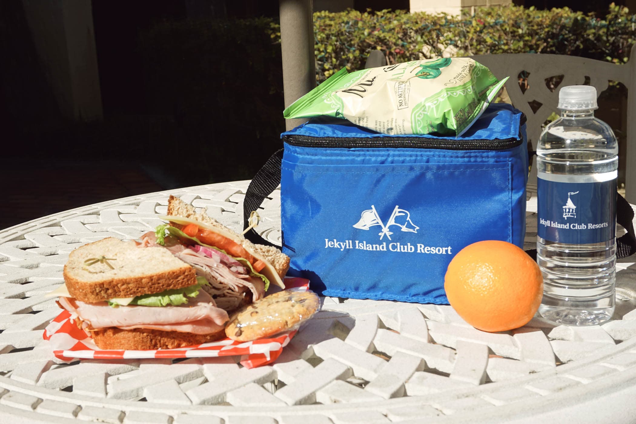 A sandwich, orange and a bottle of water on a patio table from the Pantry